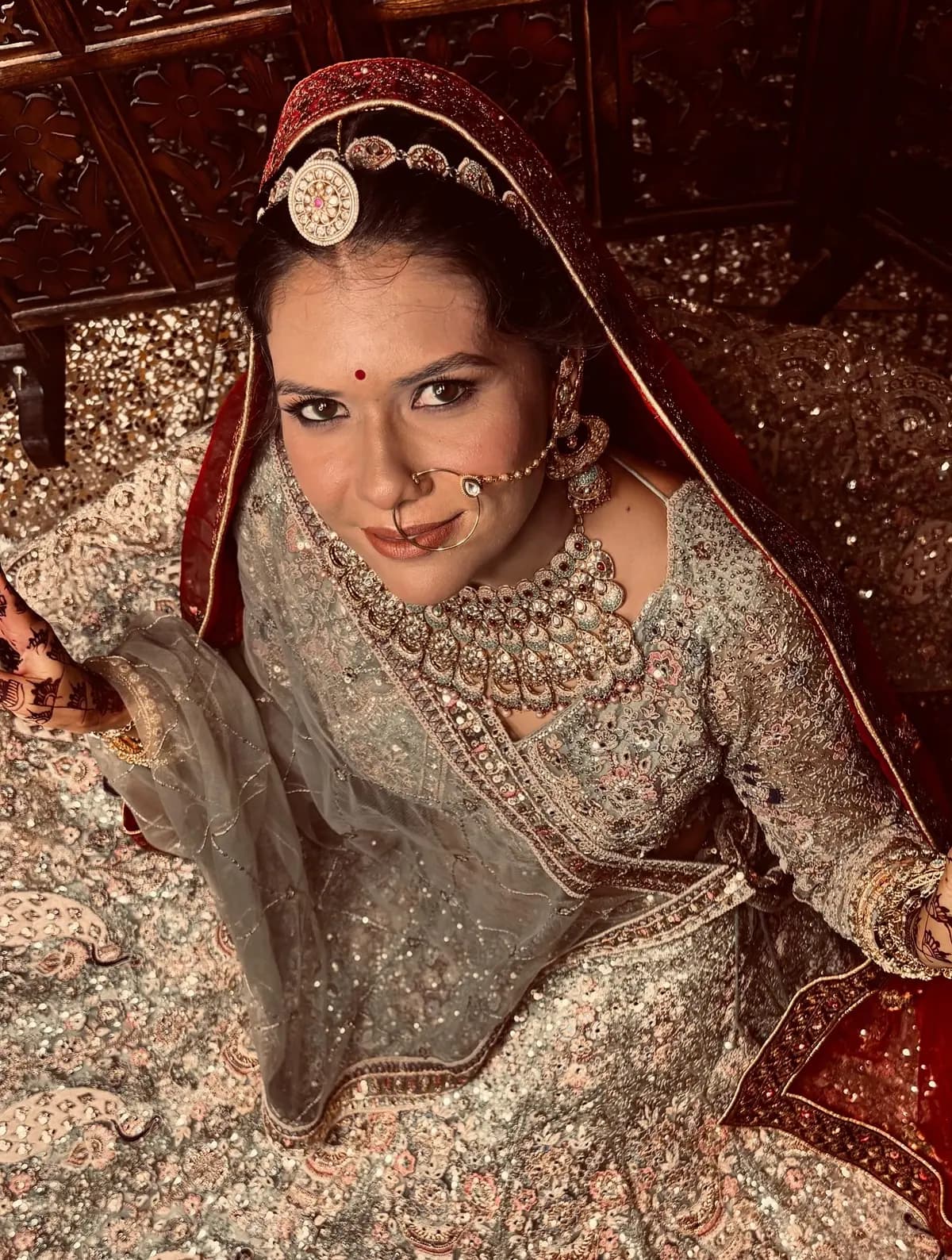 Bride in an embroidered ivory lehenga and red dupatta, looking up at the camera with soft glam makeup